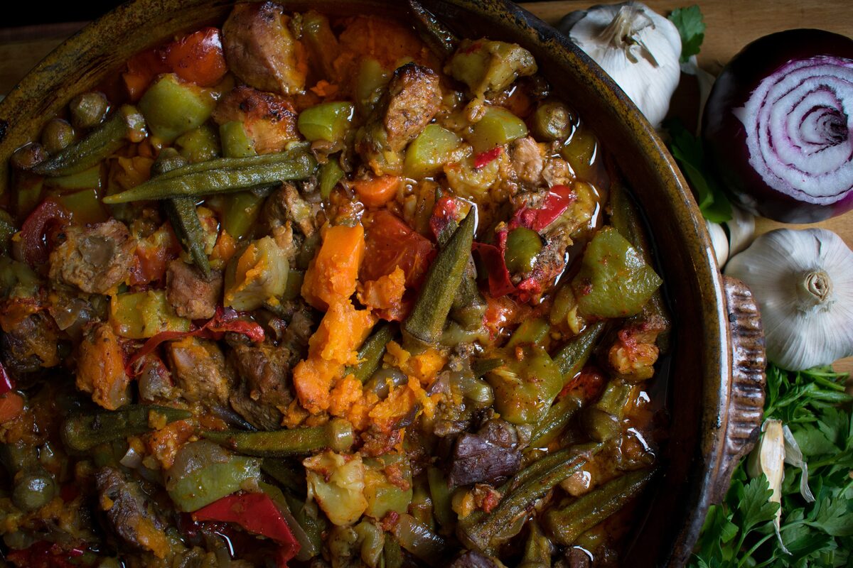 Turli tava in a clay dish, a stew of meat, okra, peppers and carrots, with garlic and an onion beside it