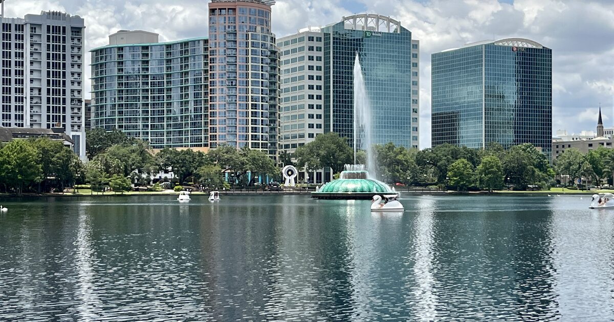 Downtown Orlando high-rises reflected in Lake Eola, with the fountain and paddle boats in front under a cloudy sky