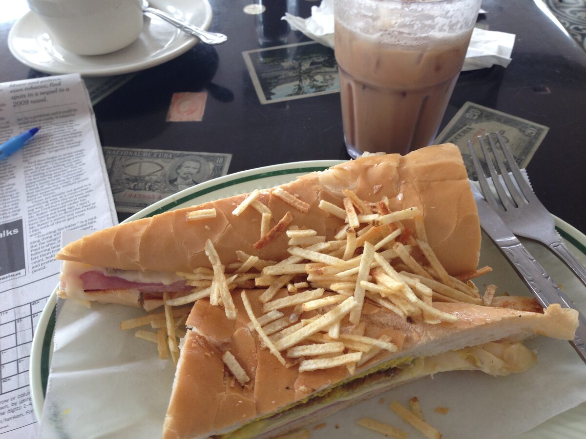 A pressed Cuban sandwich cut in half on a plate with potato sticks, next to an iced coffee