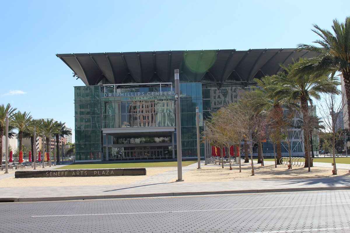 The glass-fronted Dr. Phillips Center with its sweeping dark roof, seen across Seneff Arts Plaza and palm trees