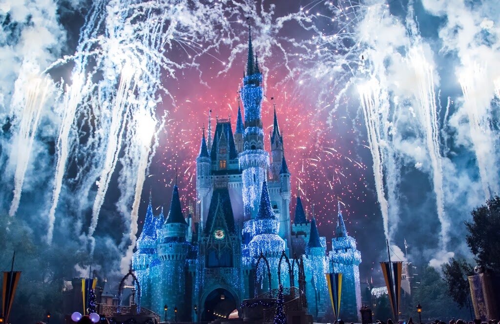 Fireworks bursting and white sparks streaming up behind the lit blue Cinderella Castle at night