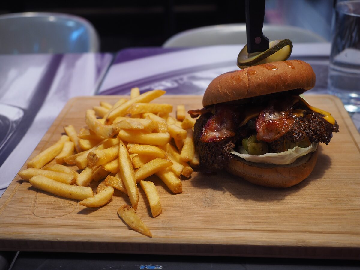 A bacon cheeseburger with a pickle skewer next to a pile of fries on a wooden board