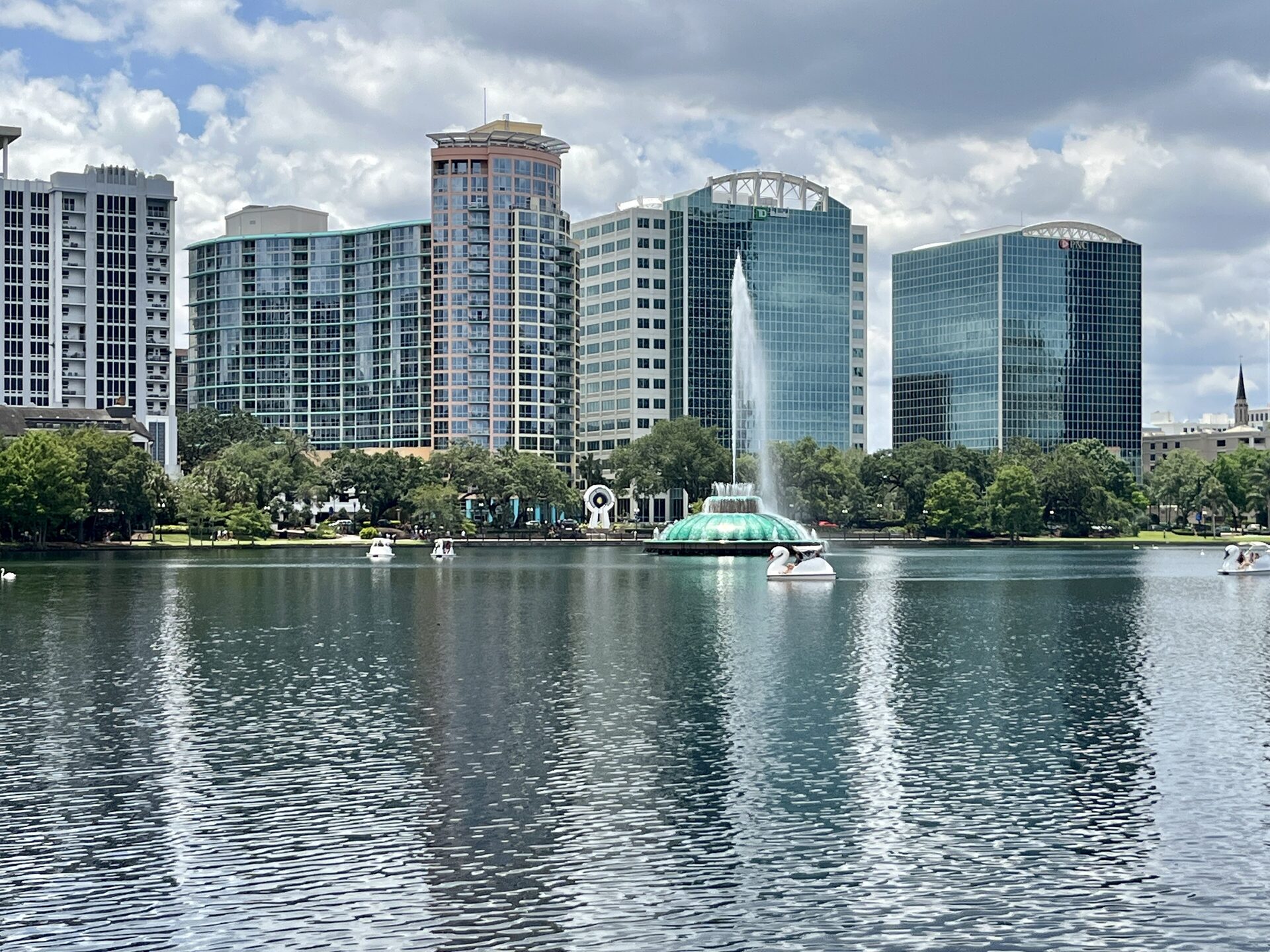 Downtown Orlando high-rises reflected in Lake Eola, with the fountain and paddle boats in front under a cloudy sky