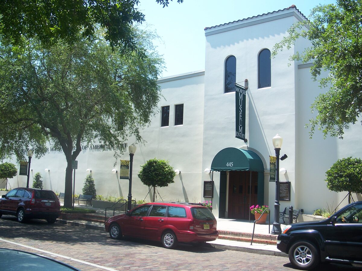 The white stucco entrance of the Morse Museum with a green awning, arched windows and a brick street in front