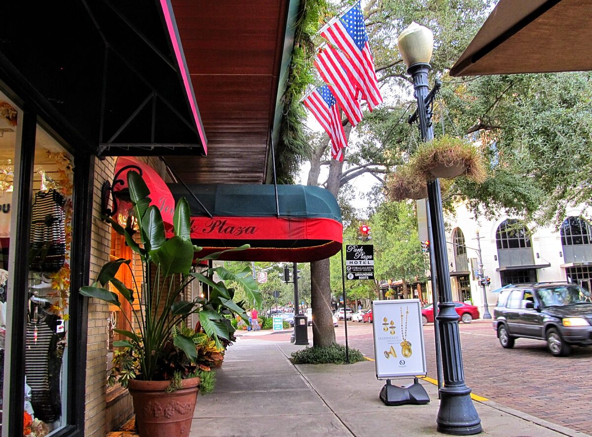 Shop awnings, an American flag and hanging baskets along a brick-paved stretch of Park Avenue under live oaks