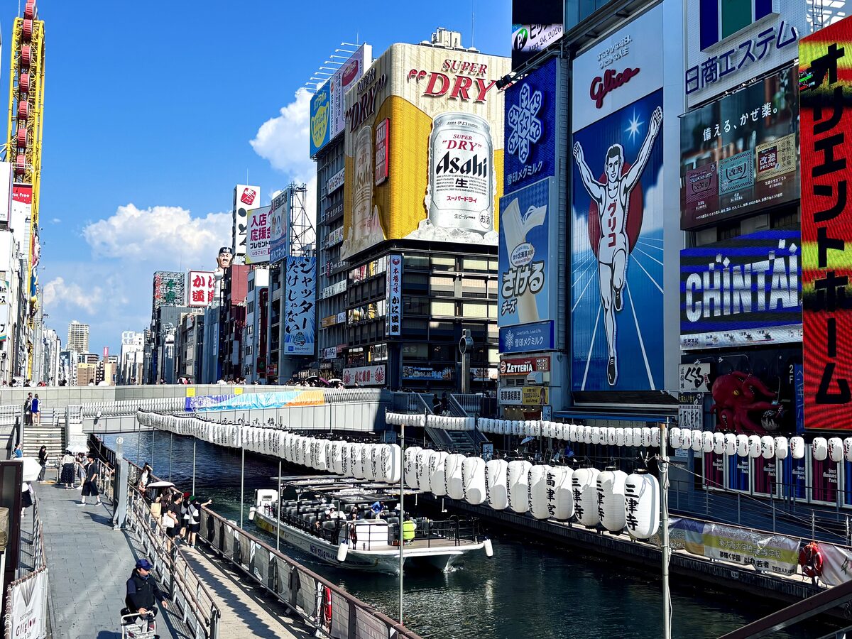 Dotonbori canal in daylight, with the Glico runner billboard above the shops and lanterns hanging along the water