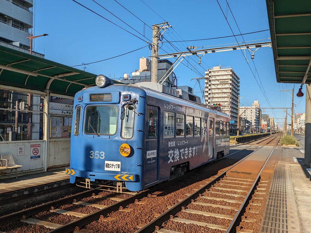 A blue Hankai tram, car number 353, stopped at a platform with overhead wires and apartment blocks behind