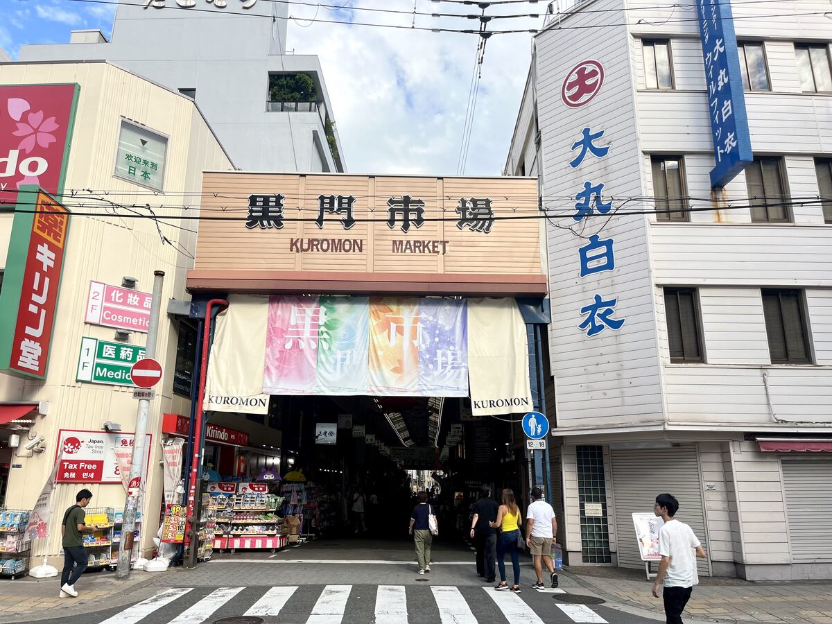 The arch of Kuromon Market at the end of its covered shopping street, with shoppers crossing the road in front of it