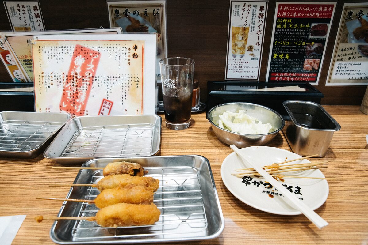 Fried kushikatsu skewers on a metal tray, with a bowl of rice and a cold drink on the table