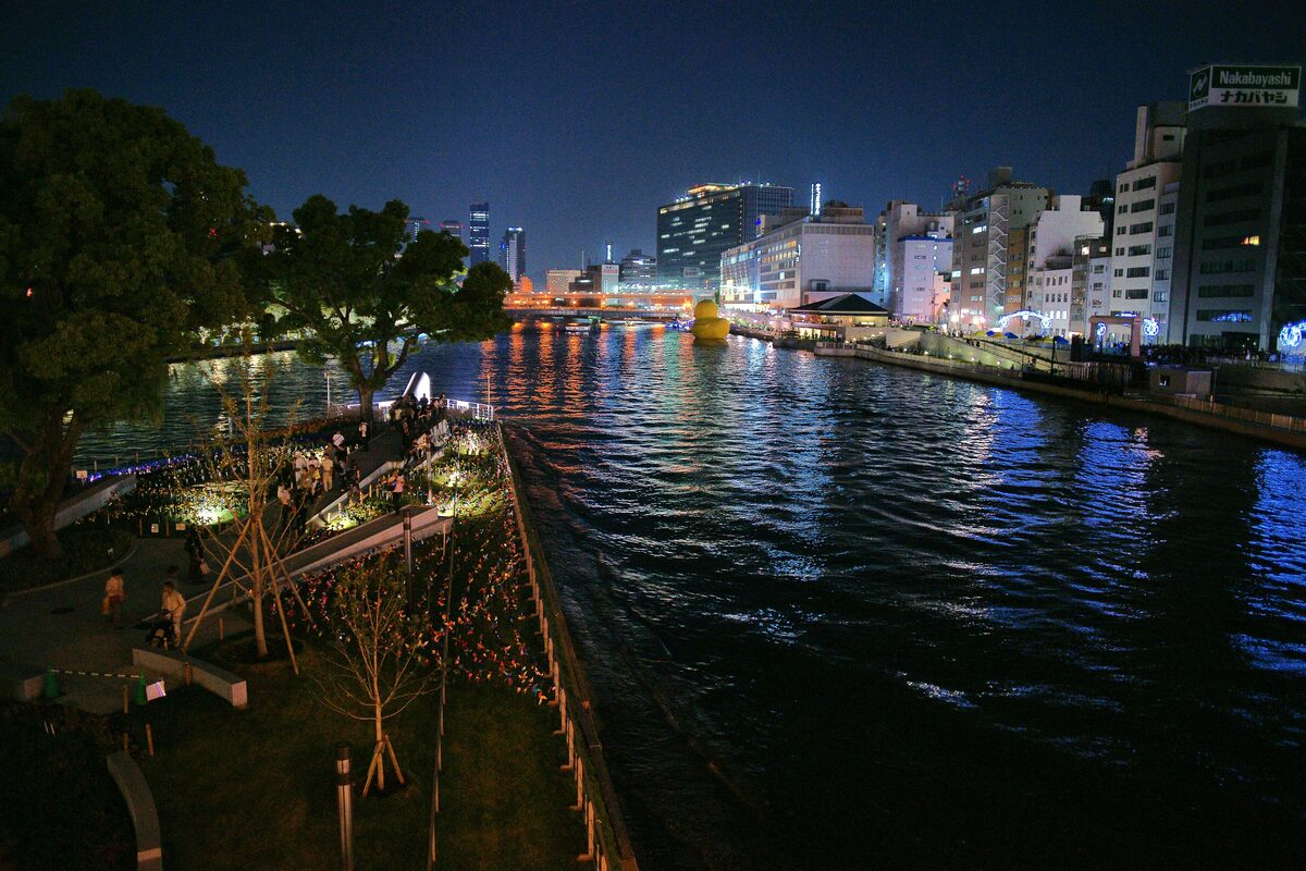 Nakanoshima park at night, with lights reflected in the river and the city towers behind the trees