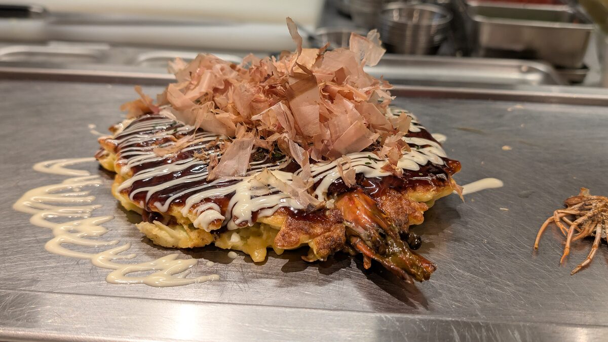 Okonomiyaki cooking on a steel griddle, topped with bonito flakes and sauce