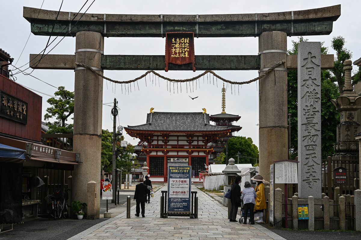 The stone torii at the entrance to Shitennoji with a red temple gate and a pagoda behind it at the end of the paved approach