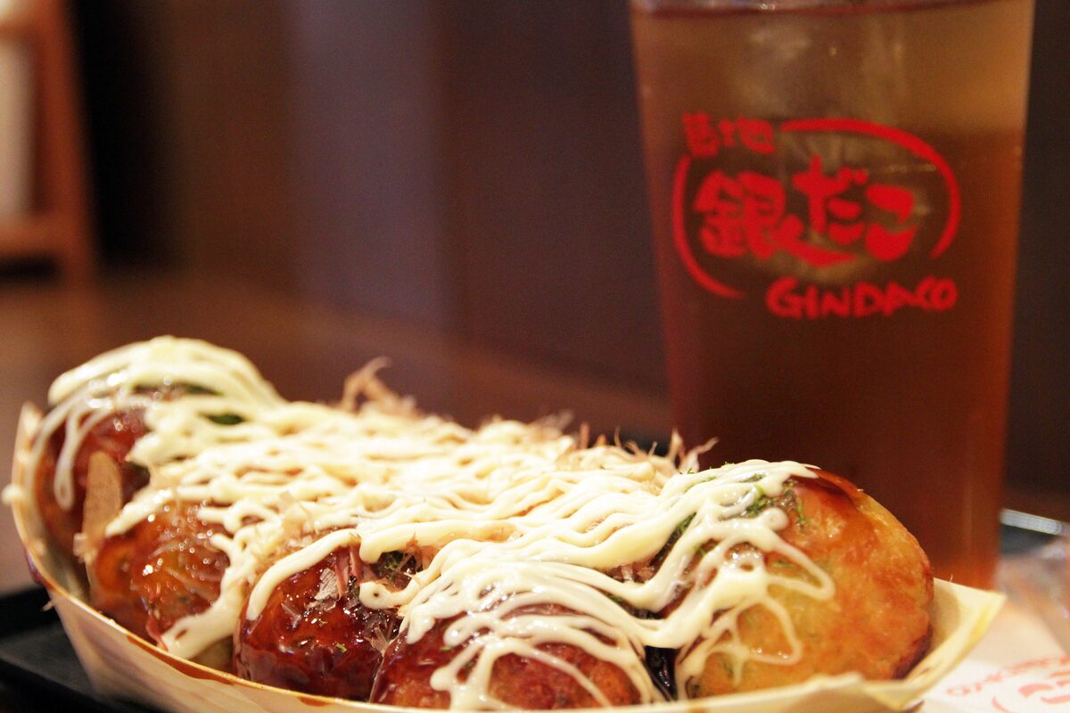 A paper boat of takoyaki balls covered in sauce, mayonnaise and bonito flakes, with an iced tea beside it