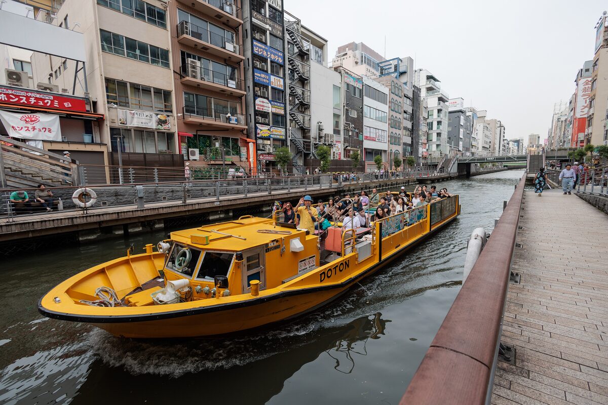 A yellow Tombori river cruise boat full of passengers passing along the Dotonbori canal between the buildings and the boardwalk