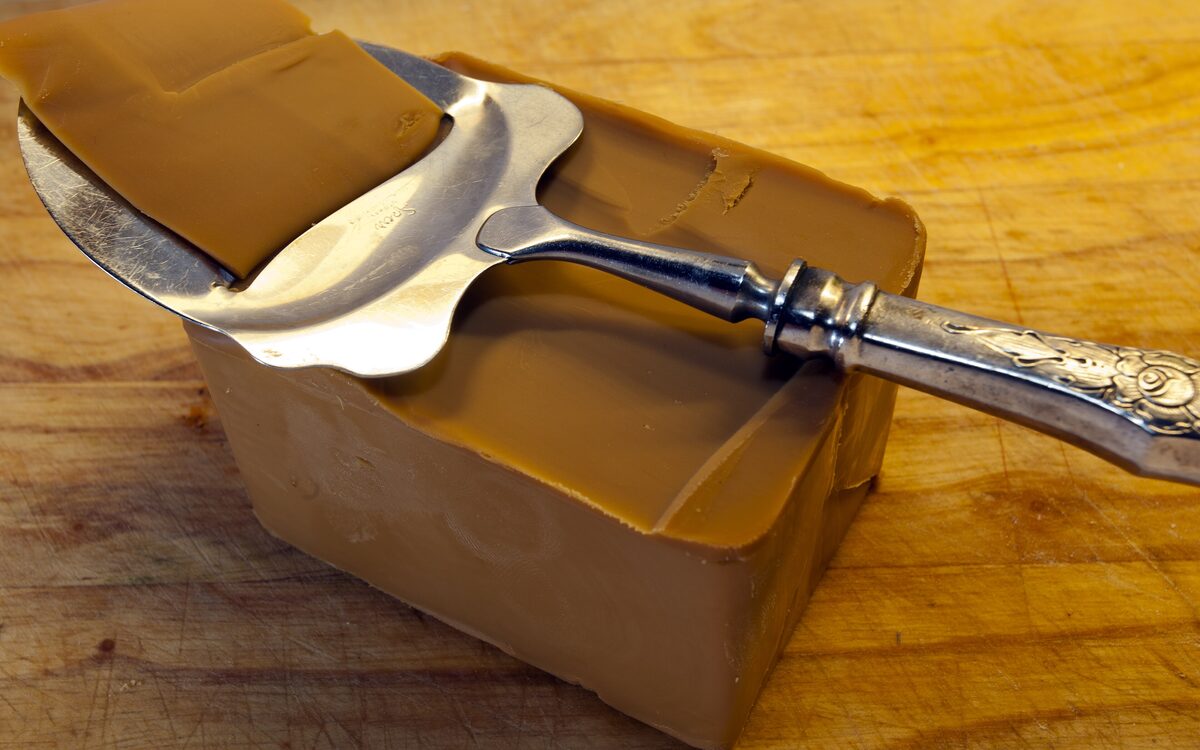 A block of brown cheese with a silver cheese slicer resting on top, on a wooden board
