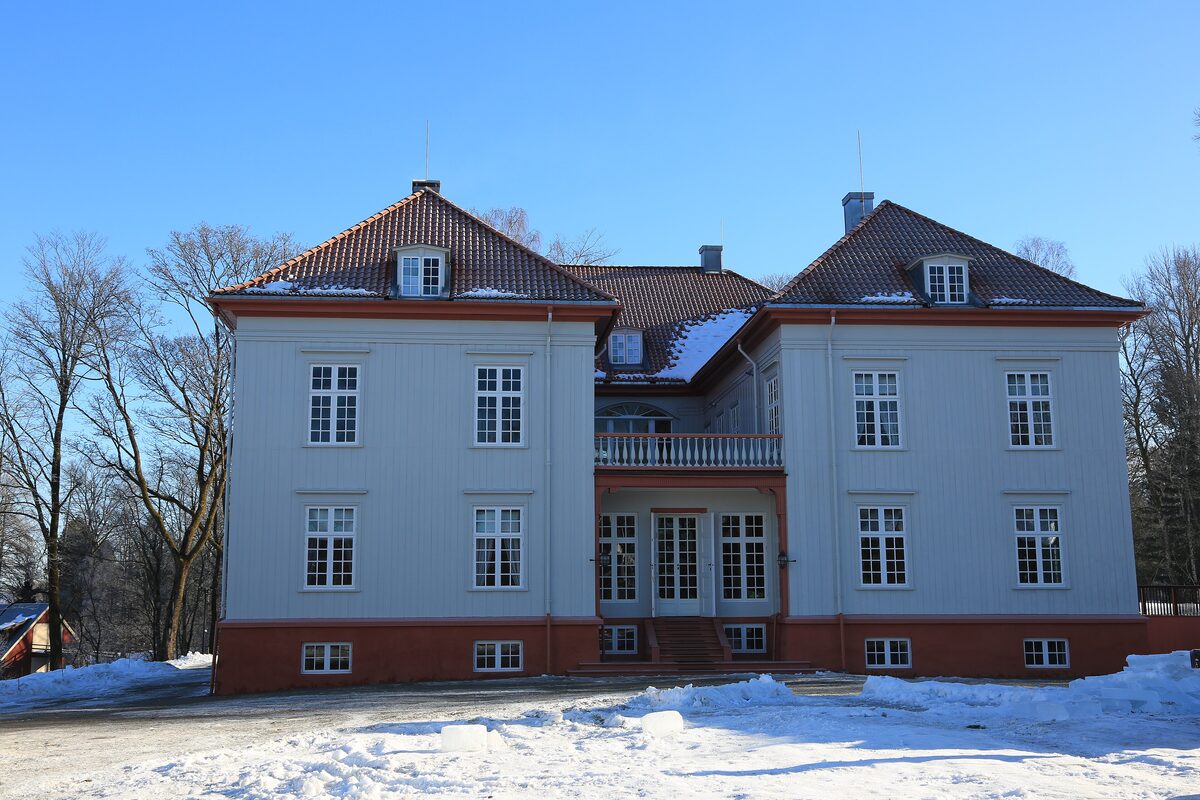 The white two-storey manor house of Eidsvollsbygningen with red trim, behind snowy grass and bare trees