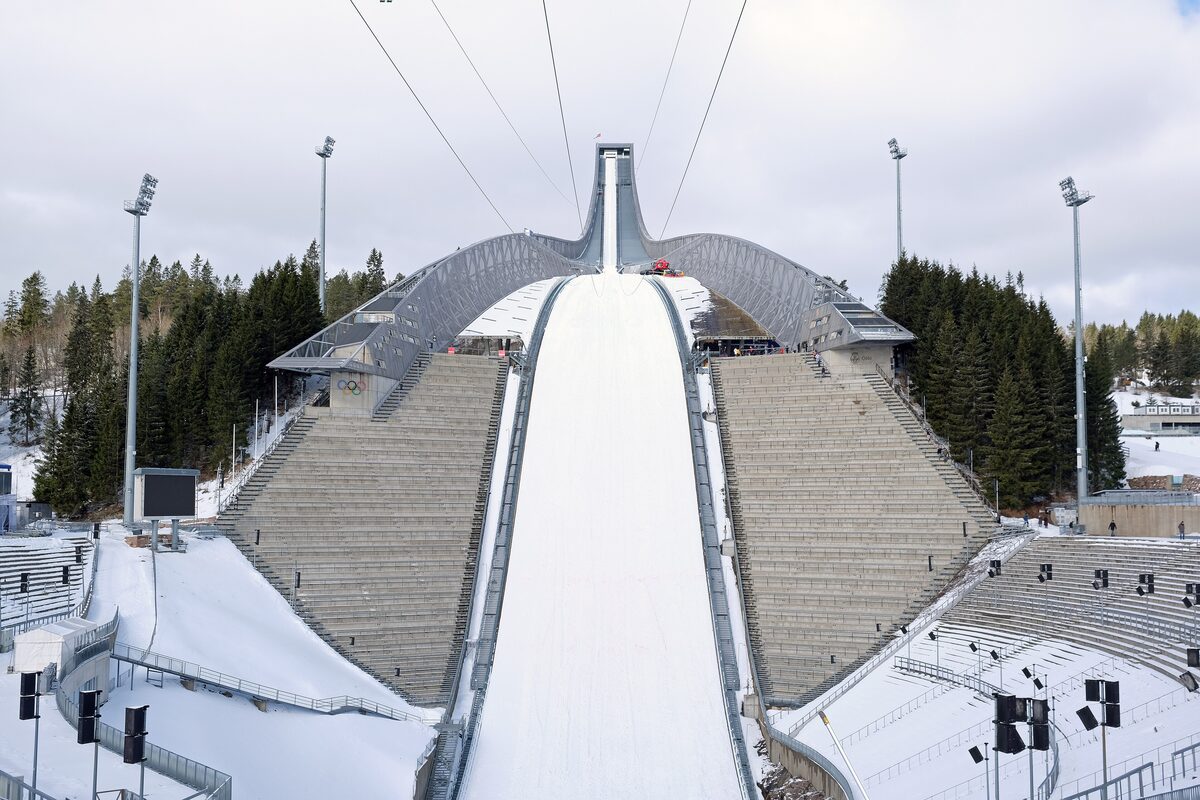 The steep white inrun of the Holmenkollen ski jump seen from the top, with snowy stands below and forest around