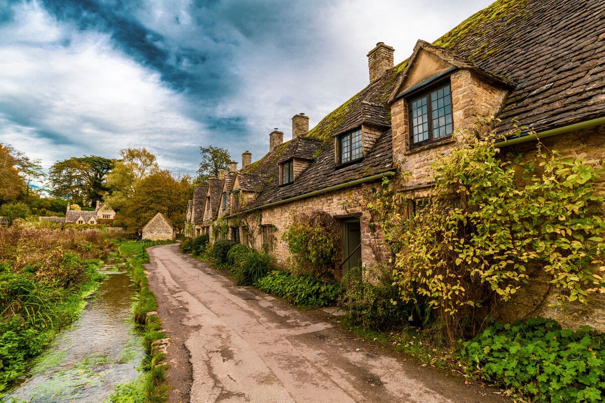 A row of old stone cottages beside a lane and a stream in the Cotswold village of Bibury