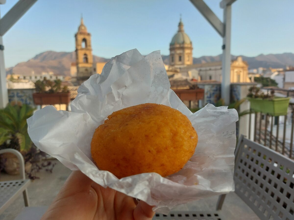 An arancina held up on paper, with Palermo's church domes and hills behind