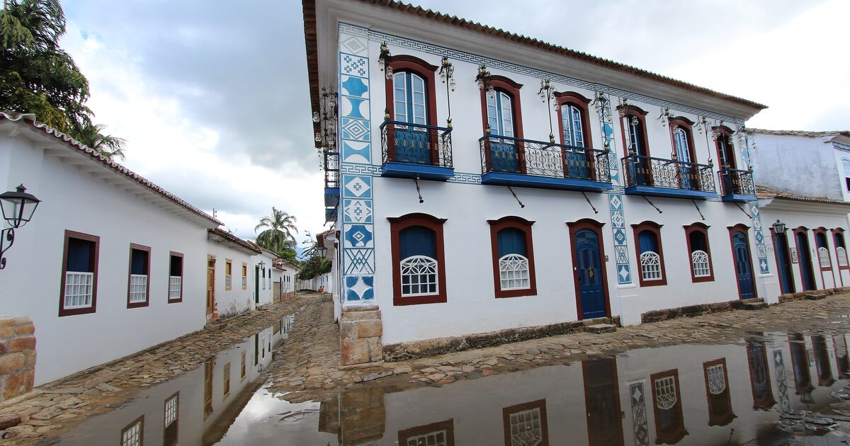 A whitewashed colonial house with blue balconies and window frames, mirrored in the tide water on a Paraty street