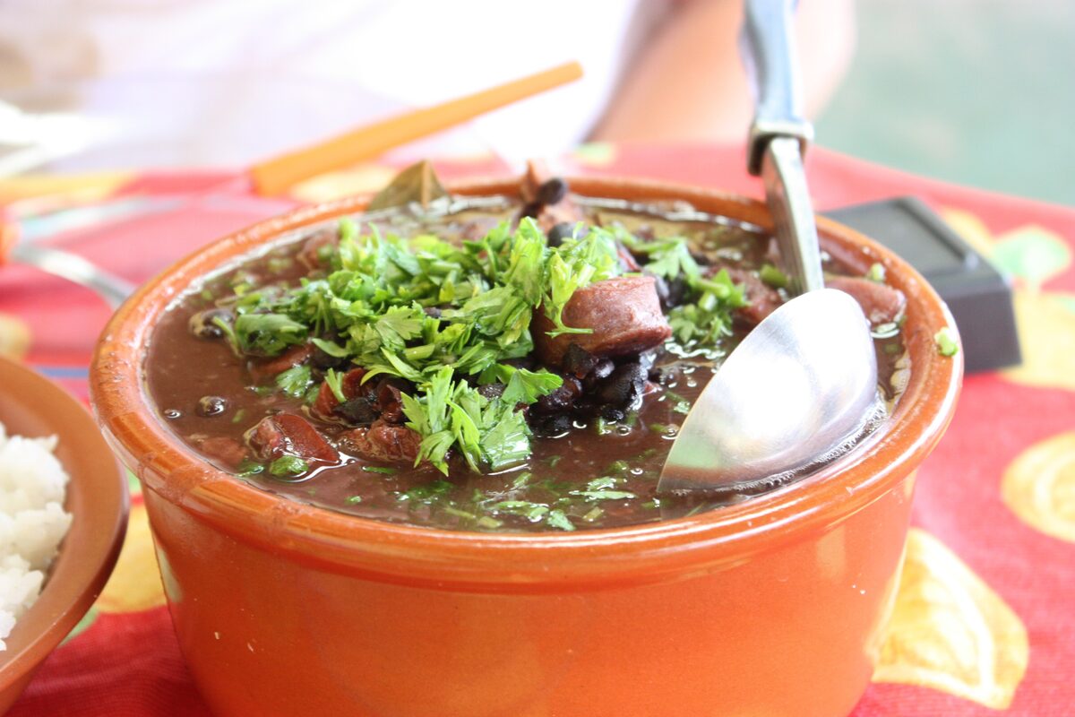 A clay pot of feijoada, black beans and pork stewed dark and topped with chopped parsley, with a spoon in it