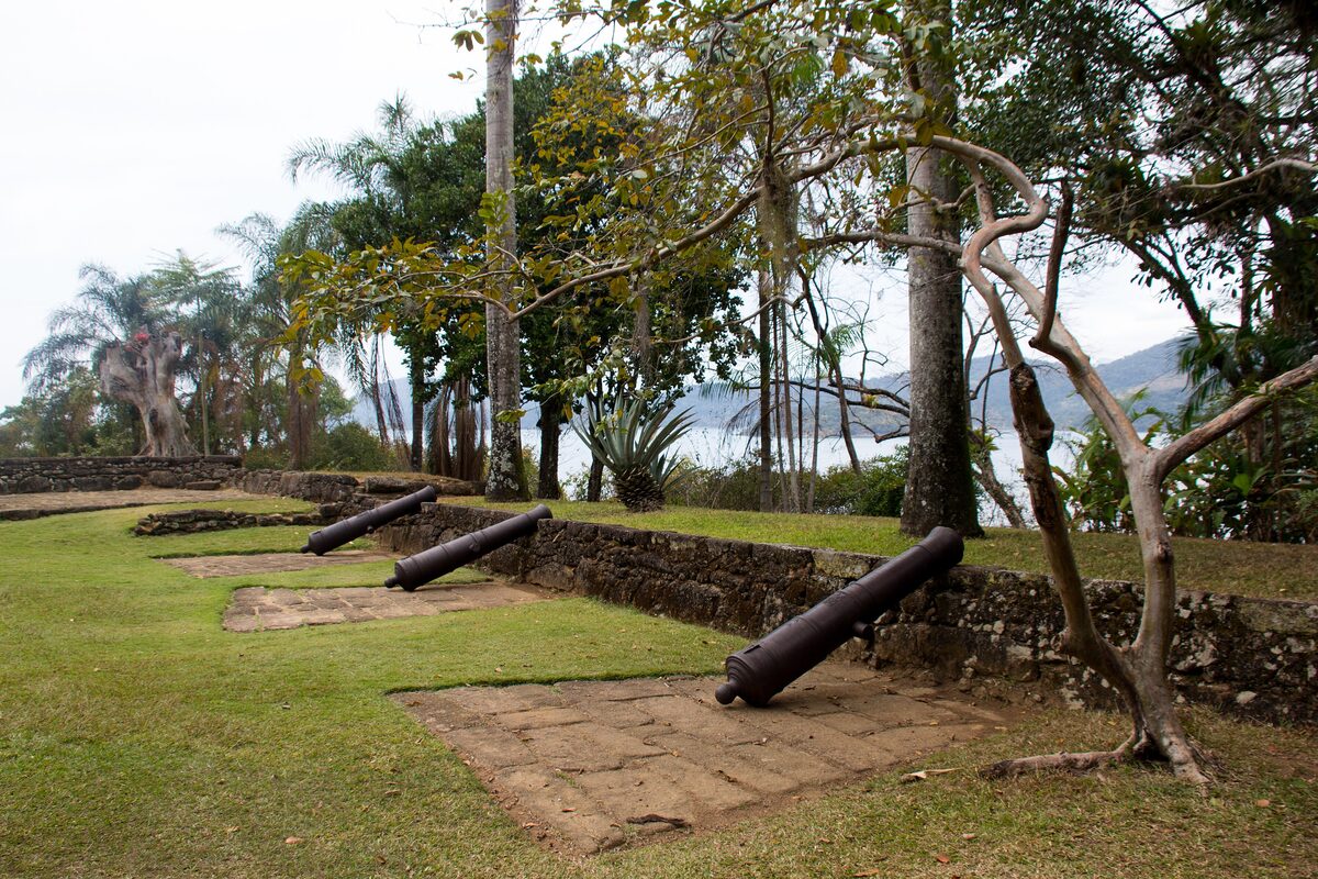 Old iron cannons on a grassy bastion behind a low stone wall at the Forte Defensor Perpétuo, with palms and the bay beyond