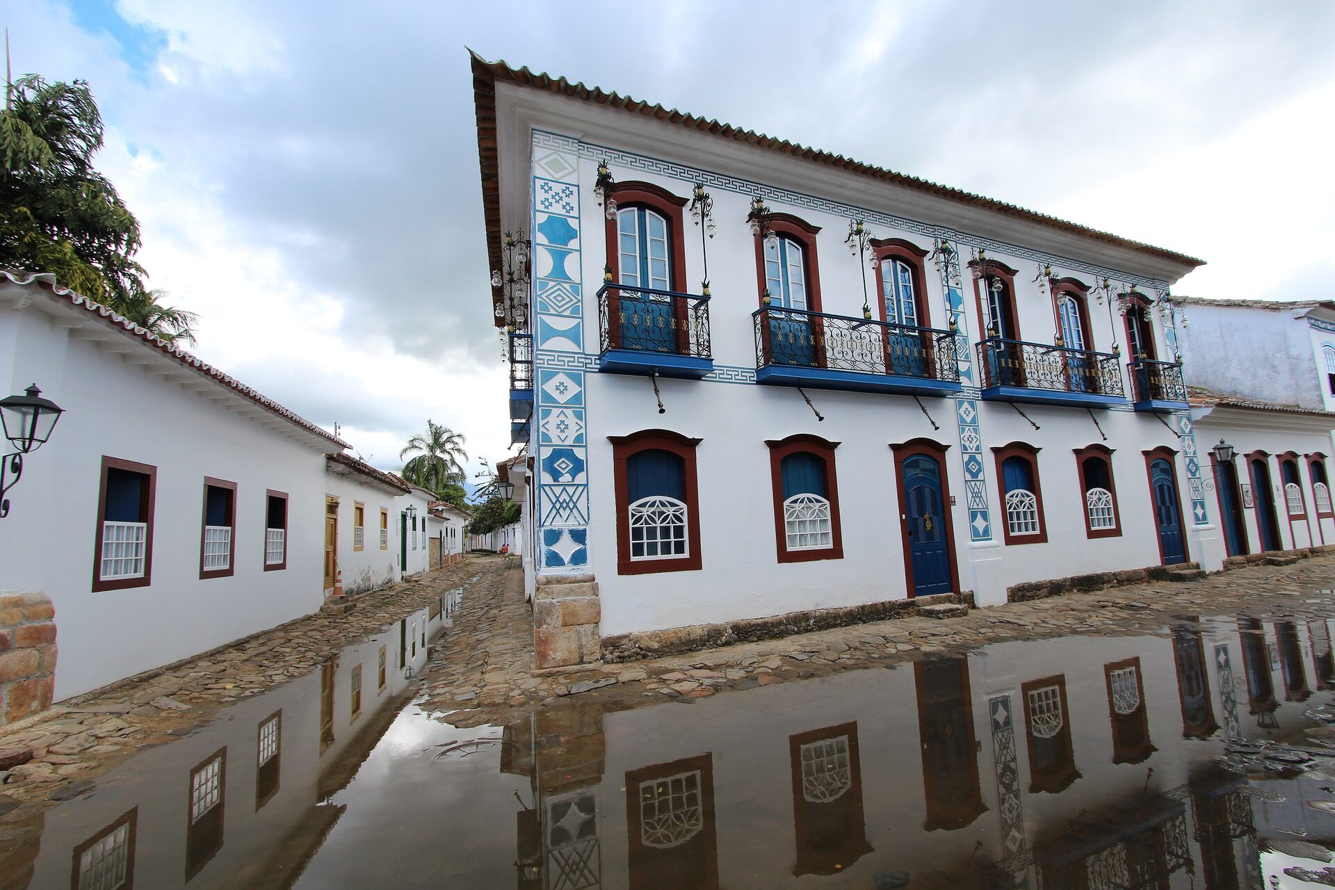 A whitewashed colonial house with blue balconies and window frames, mirrored in the tide water on a Paraty street