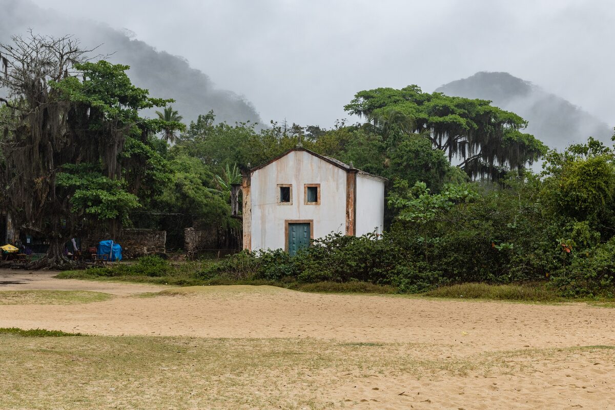 A small white chapel beside a wide sandy beach at Paraty-Mirim, under low cloud and forest