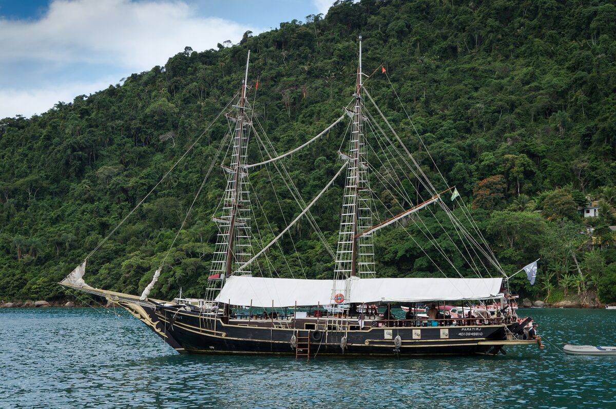 A two-masted wooden schooner on the green water of Paraty bay, with forested hills behind