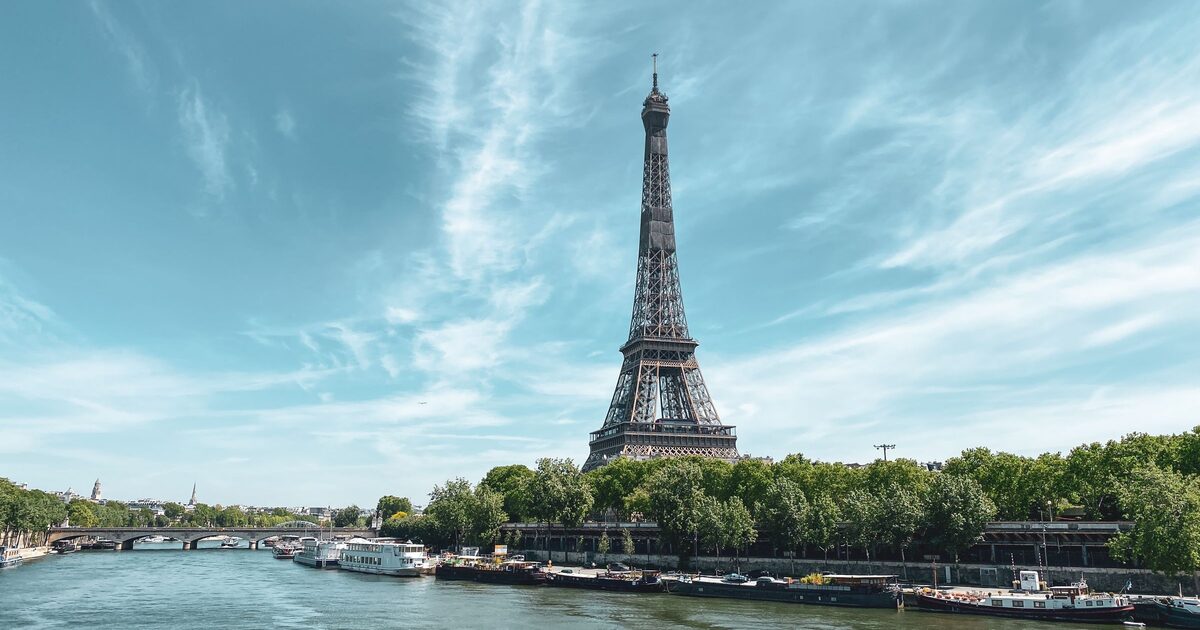The Eiffel Tower rising above the trees on the bank of the Seine, with moored boats and a bridge under a pale blue sky