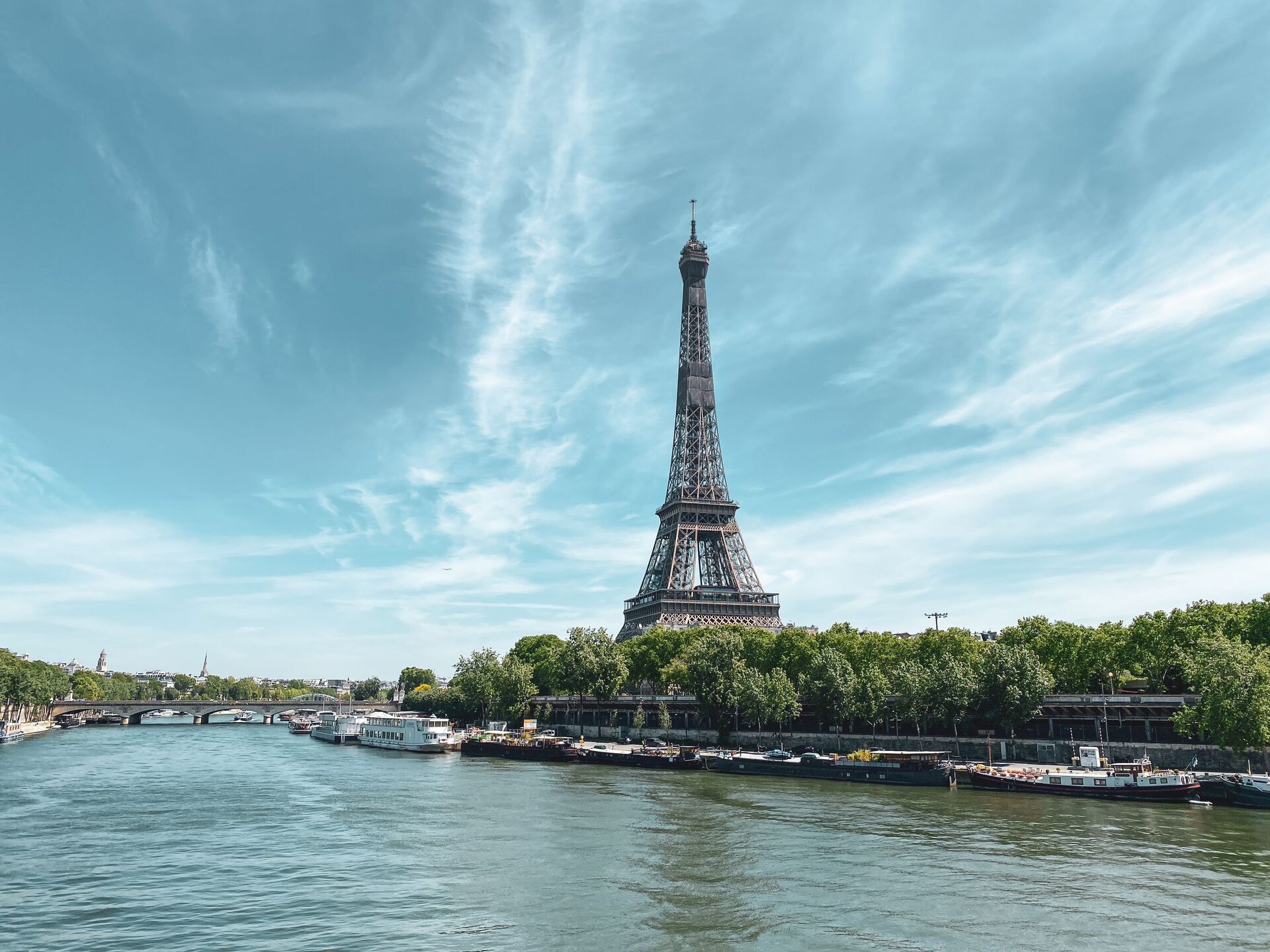 The Eiffel Tower rising above the trees on the bank of the Seine, with moored boats and a bridge under a pale blue sky