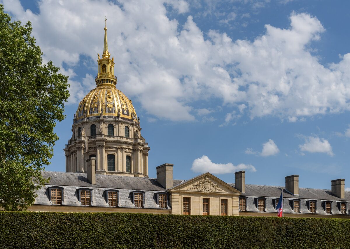 The gilded dome of Les Invalides rising behind a clipped hedge and the grey roofs of the hospital wing, under a blue sky with white clouds