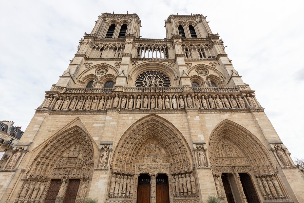 The west front of Notre-Dame seen from the parvis, with the twin towers, the rose window and the three carved portals under a cloudy sky