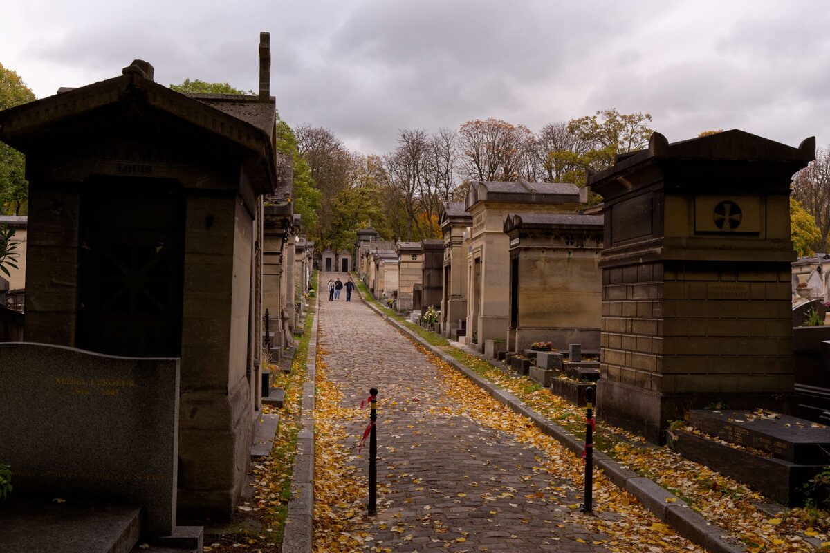 A cobbled path between rows of stone family tombs and chapels in Père Lachaise Cemetery, strewn with autumn leaves