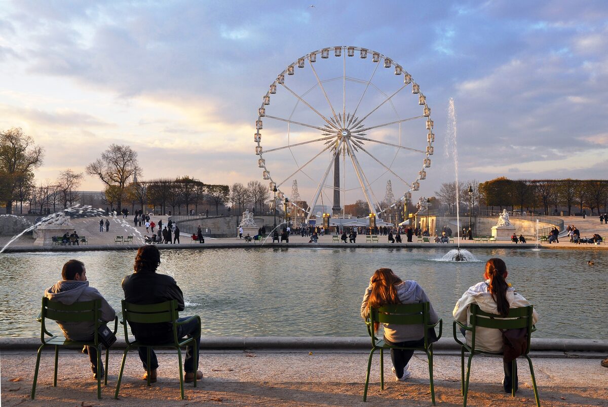 Four people in green metal chairs beside the octagonal basin in the Tuileries Garden at sunset, with the Ferris wheel behind
