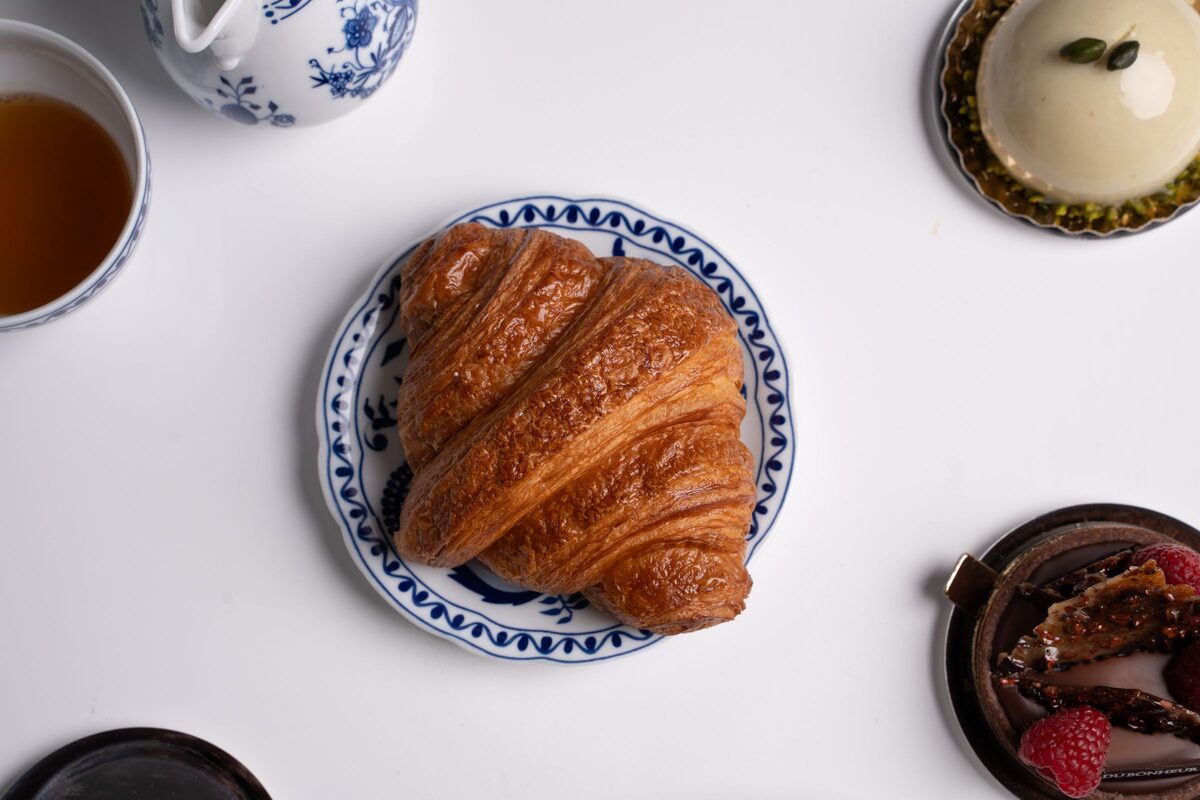 A golden croissant on a blue-and-white plate, seen from above, with tea and small cakes around it