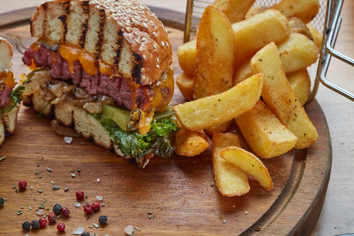 A thick cheeseburger cut in half beside a pile of potato wedges on a wooden board