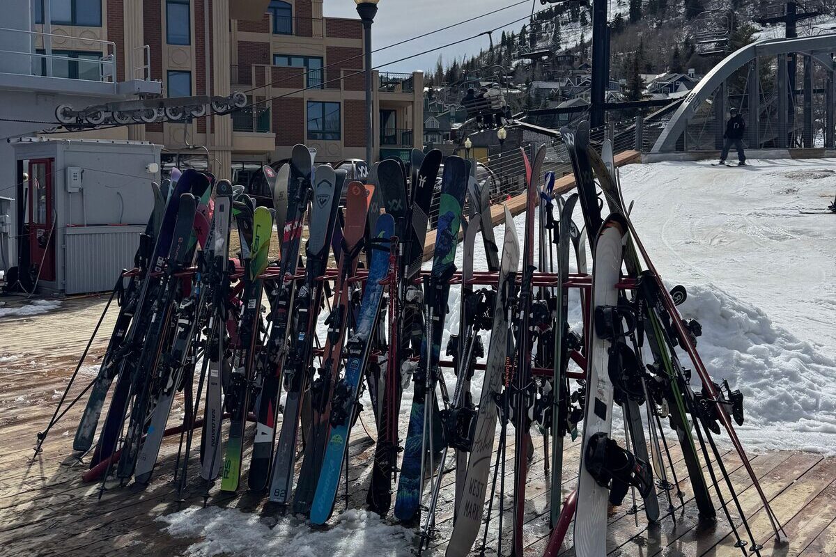 A rack of skis and poles on a snowy plaza beside a lift in Park City, with an arched footbridge and the hillside behind