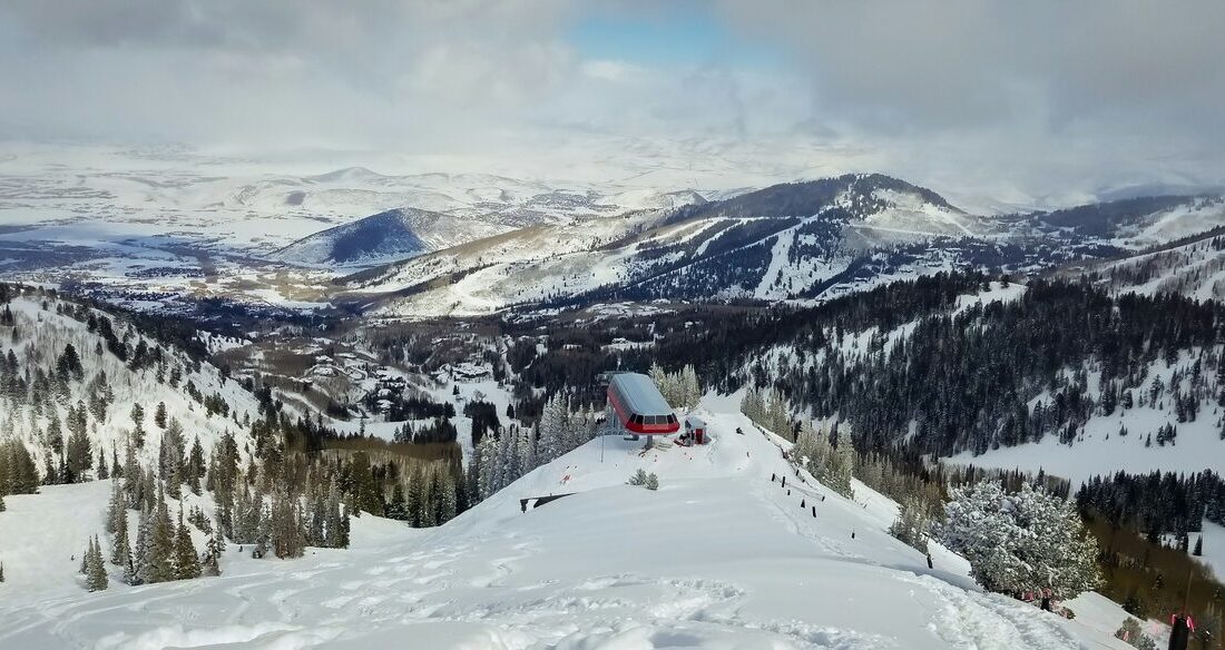 Looking down on the top of the Ninety-Nine 90 lift at 9,990 feet, a red lift building on a snowy ridge with the valley and distant ranges beyond