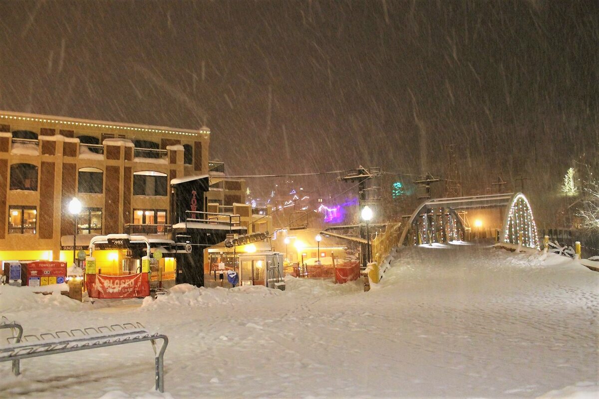 Snow falling on the Town Lift plaza in Old Town Park City at night, with the lit bridge and Main Street buildings
