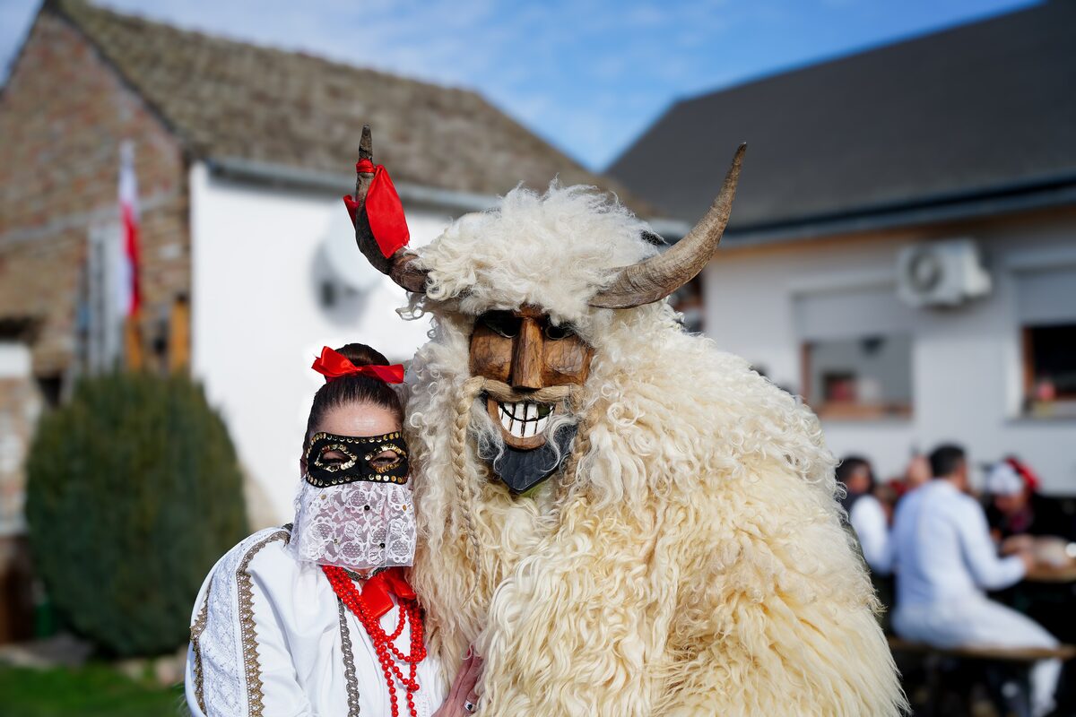A busó in a sheepskin coat and a carved wooden mask with horns, standing next to a woman in a lace veil and folk dress
