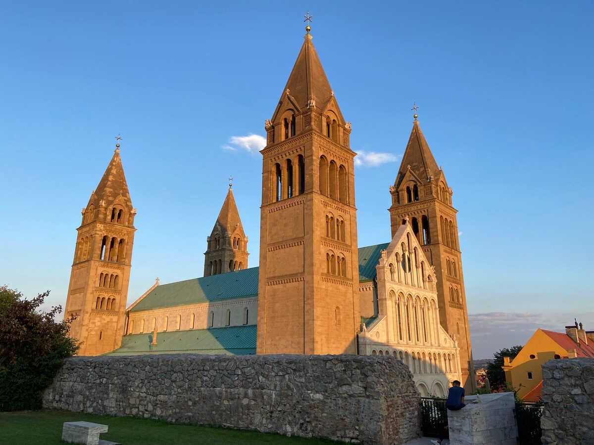 Pécs Cathedral with its four tall towers glowing in the evening sun against a blue sky, behind an old stone wall