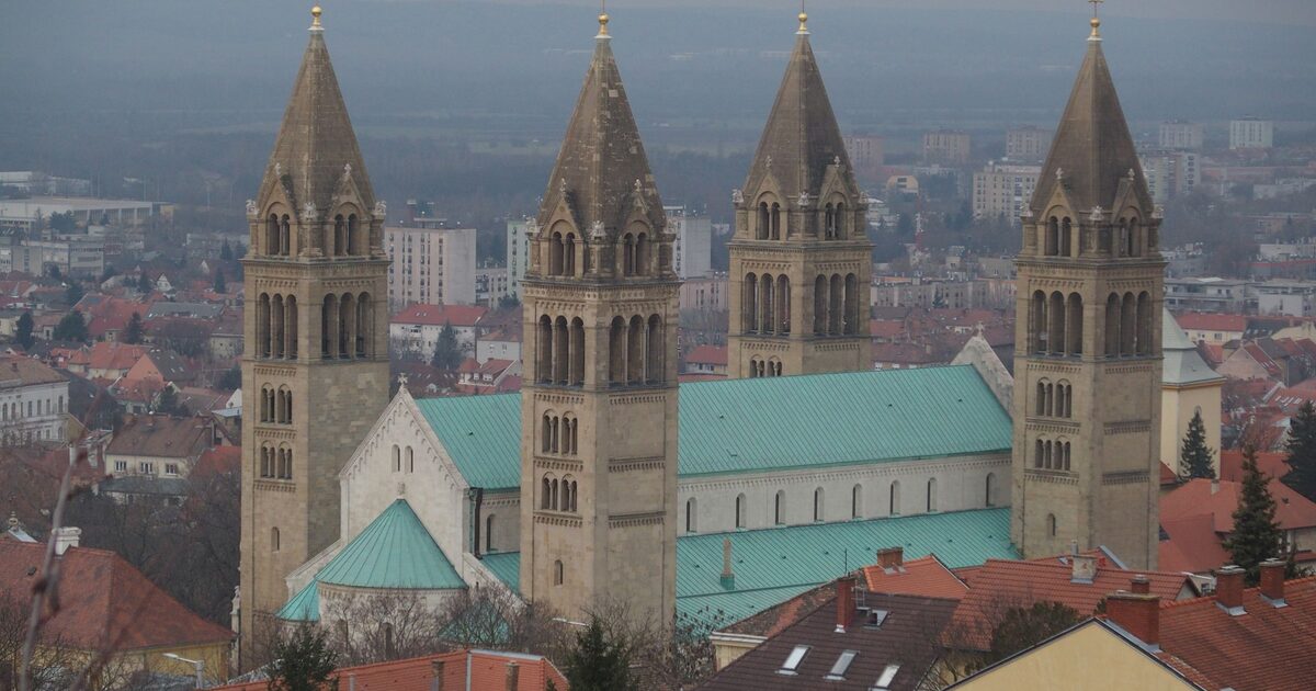 The four towers and green roofs of Pécs Cathedral above the rooftops of the city, with housing blocks and the plain stretching away behind
