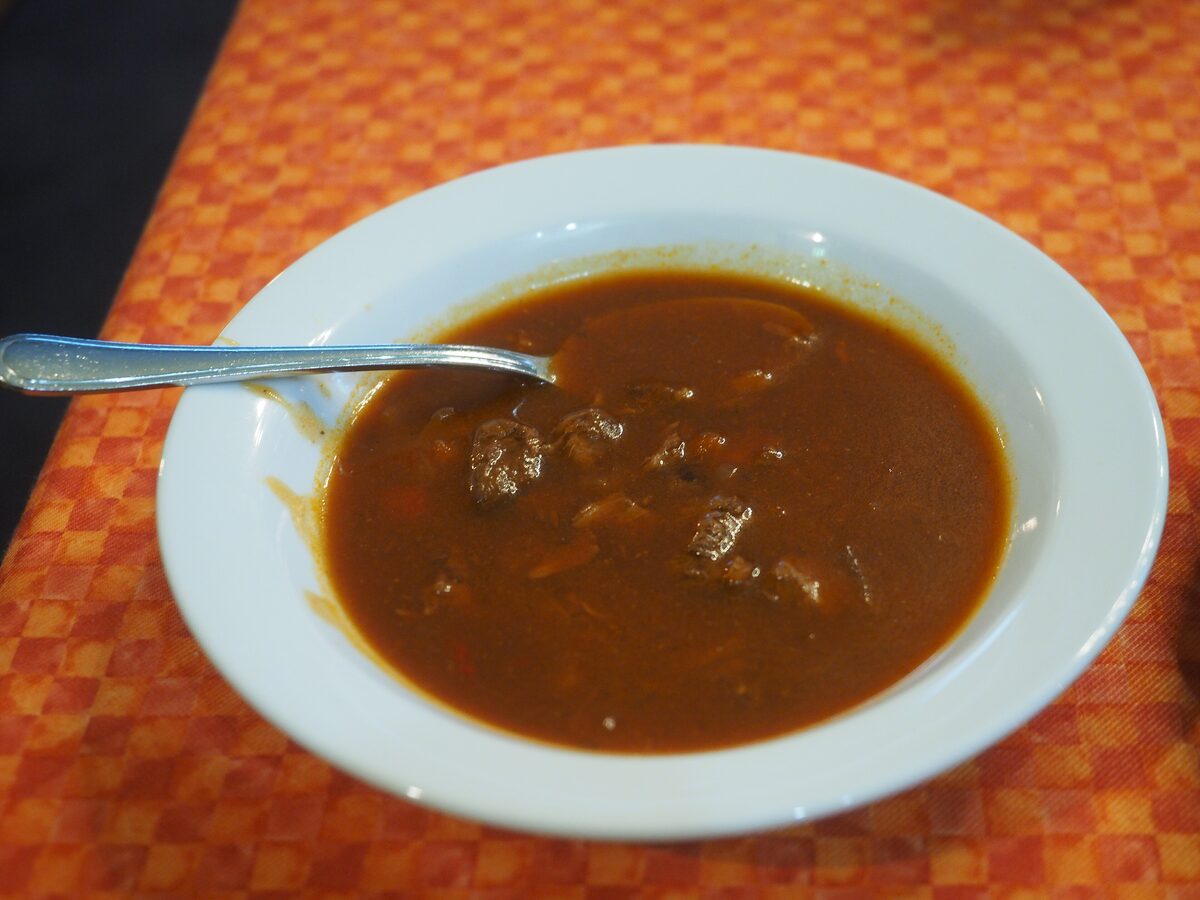 A bowl of goulash soup with chunks of beef in a red paprika broth on an orange tablecloth