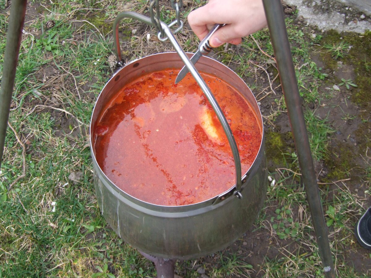 A cauldron of bright red halászlé fish soup hanging over an outdoor fire, with a ladle in it