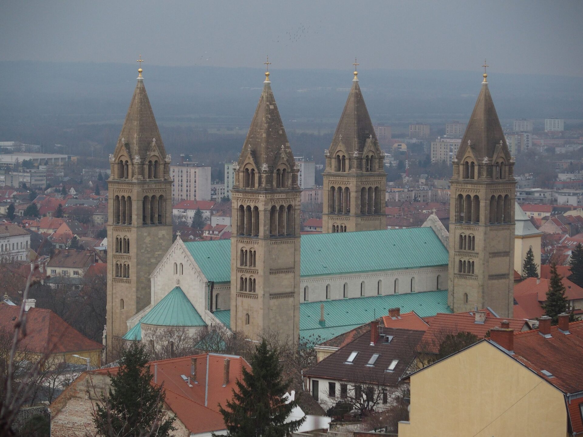 The four towers and green roofs of Pécs Cathedral above the rooftops of the city, with housing blocks and the plain stretching away behind