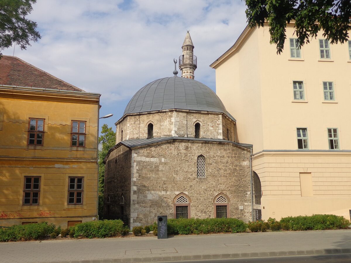 The Jakovali Hassan Mosque in Pécs, a stone building with a grey dome and a slim minaret, between two yellow houses