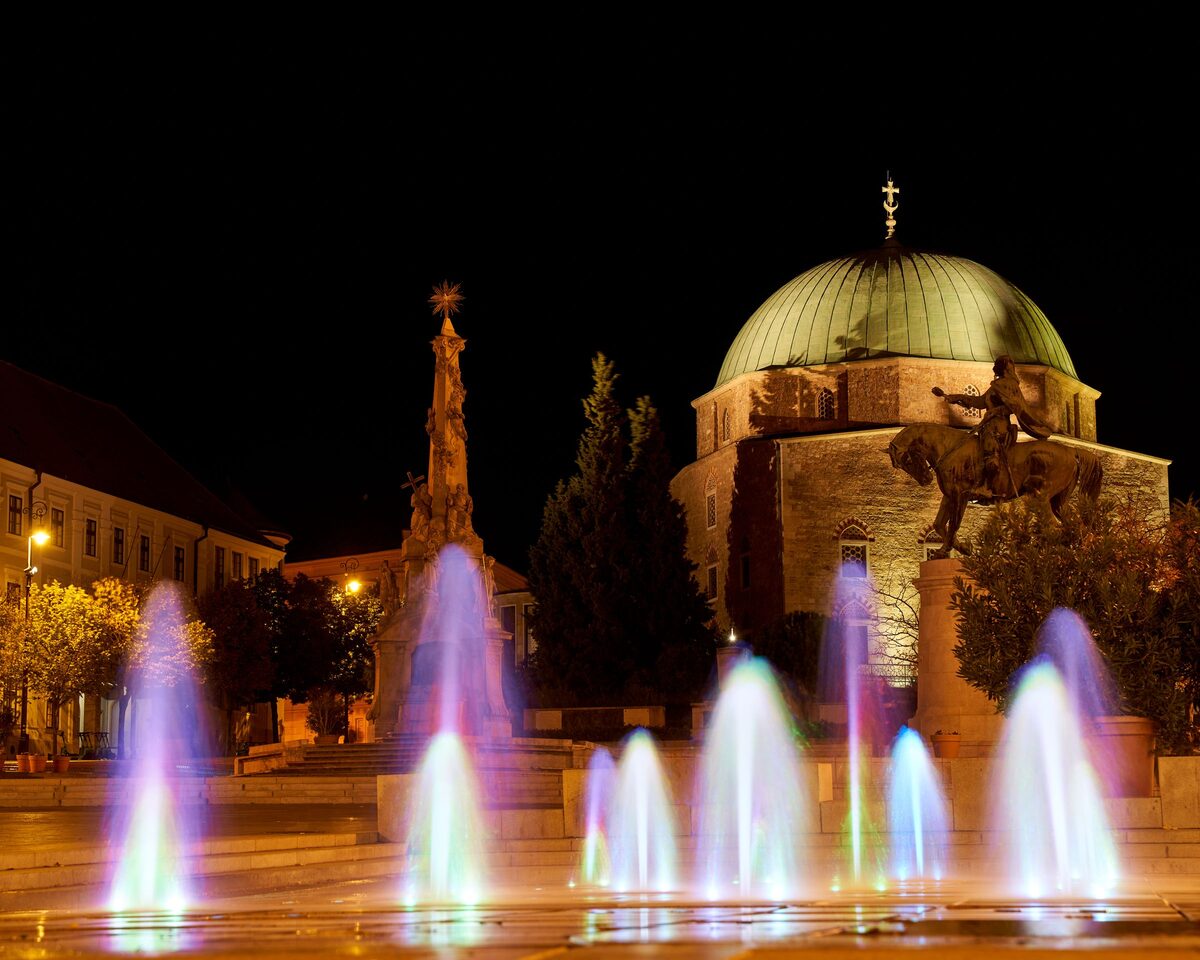 The green dome of the Mosque of Pasha Qasim at night, with colored fountains and the Holy Trinity column in front