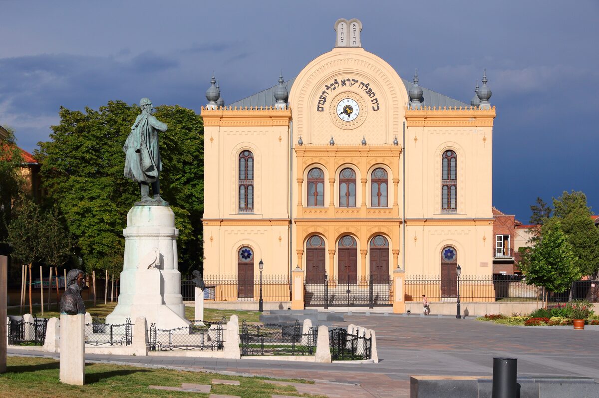 The yellow facade of the Pécs synagogue with arched windows and a round window on top, with a statue on a pedestal in the square in front