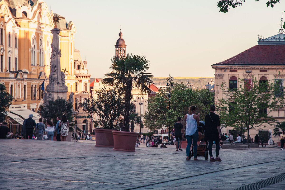 Széchenyi Square in Pécs in the evening light, with the Holy Trinity column, a palm tree in a pot and people walking past ornate buildings